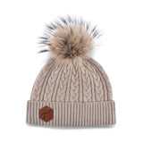 Women's beige, cashmere, knitted, beanie with light brown fur pom top and brown, leather, hexagonal patch on the brim with debossed T3 logo.